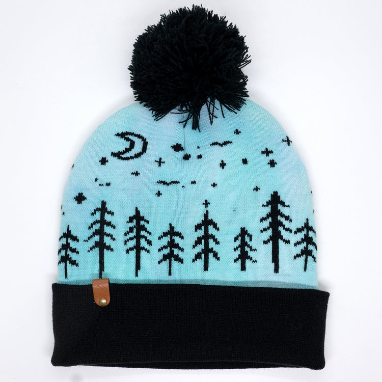 A light blue and black knit beanie hat with a black pom-pom on top, featuring a black knit pattern of stars, a crescent moon, and pine trees on a light blue background, with a black ribbed cuff and a small brown leather accent.