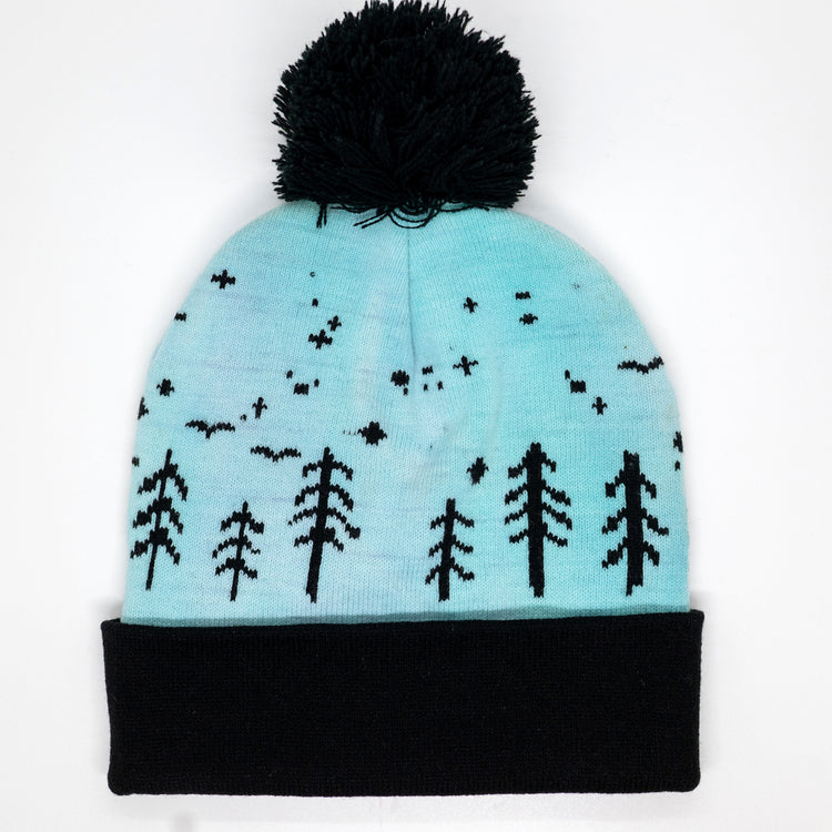 A light blue knit beanie with a black pom-pom and black cuff, featuring a knitted pattern of black trees and stars on the front.